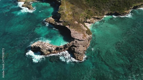 Aerial view of stunning rocky shoreline with waves pounding cliffs under clear, sunny sky, with water so clear it's turquoise. Drone is moving along a circular trajectory.