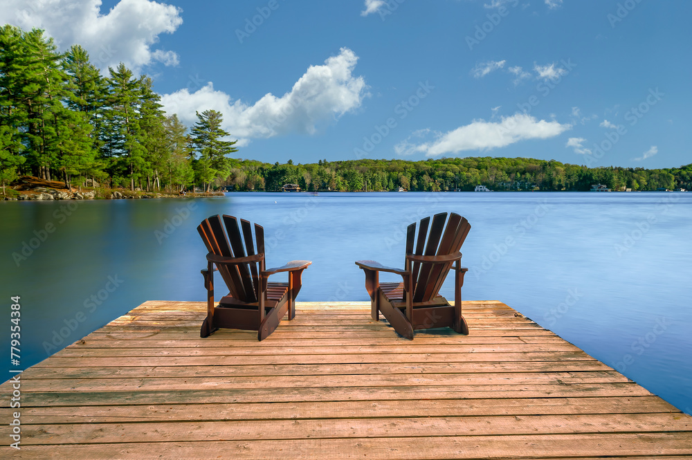 Two Adirondack chairs sit on a wooden dock beside a calm lake. Rustic cottages nestled among lush trees dot the shoreline, completing the tranquil scene.