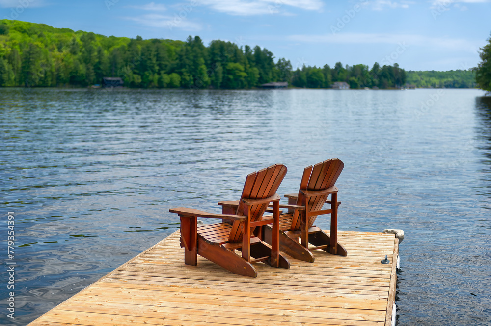 Fototapeta premium Two Adirondack chairs rest on a wooden dock, soaking up the peaceful warmth of a summer morning. Across the lake, cottages peek through the trees, completing the tranquil scene.