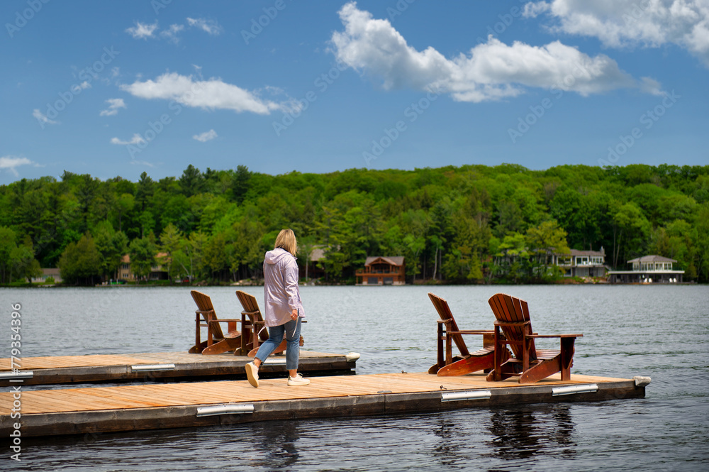 Sunny Muskoka Morning: A young woman strolls toward two Adirondack chairs on a wooden dock, overlooking the calm lake. Rustic cottages nestled among trees complete the tranquil scene.