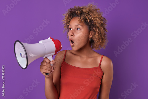 Young bold ethnic African American woman student shouting in megaphone reporting presence racial discrimination or gender inequality dressed in casual clothes stands in purple studio. Protest, strike