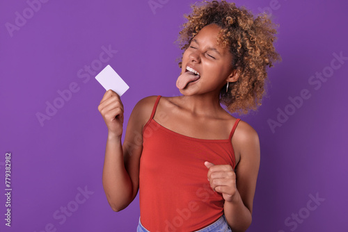 Young beautiful cheerful African American woman with credit card in hands shows tongue rejoicing at opportunity to use borrowed funds from bank to make non-cash payments stands in purple studio.