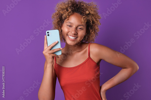 Young laughing ethnic African American woman teen with smartphone holds hand on belt and looks at camera with wide smile feeling joy due to excellent coincidence stands on purple background.