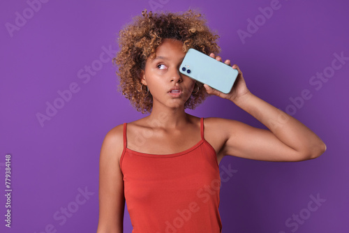 Young indecisive cute African American woman teenager closes eye with smartphone and looks up thinking about possibility of buying new phone with improved characteristics stands in purple studio.