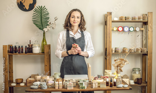 Woman soap maker in a workshop holds piece of fragrant natural soap in her hands. A lot of natural soap ready on the table, ingredients and tools on the shelves