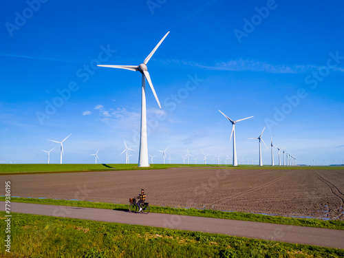 A man rides his electric bicycle down a dirt road alongside towering wind turbines on a sunny day in the Netherlands Flevoland in Spring
