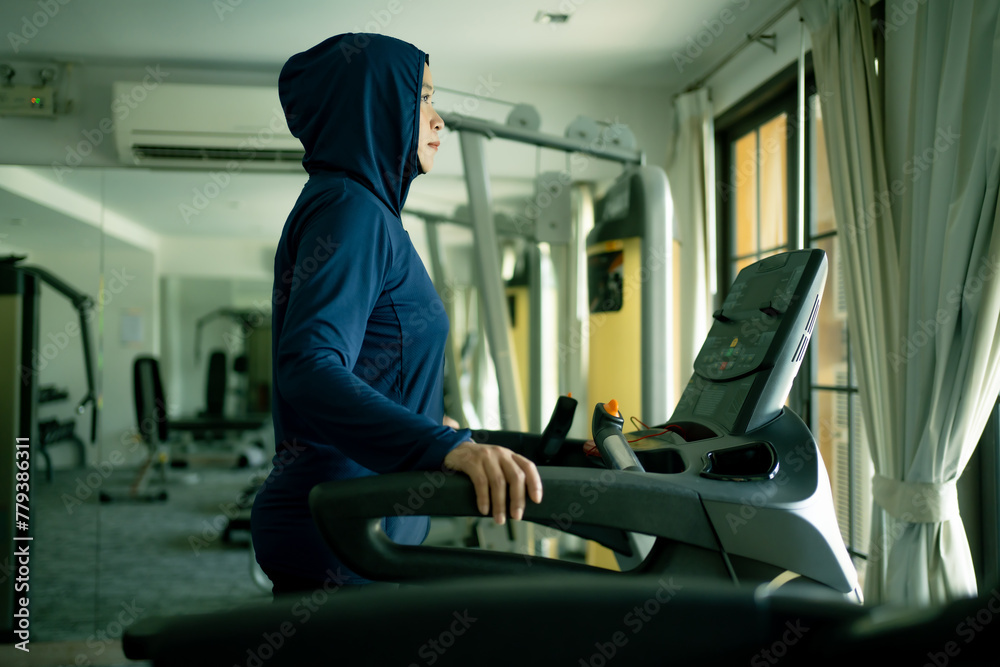 Athletic Asian Muslim Sports Woman Wearing Hijab and Sportswear Running on Treadmill. Energetic Fit Female Athlete Training in Gym Alone. Urban Business District Window View. 
concept of muslim sport.