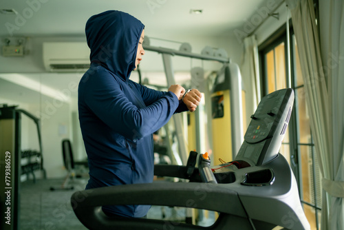 Athletic Asian Muslim Sports Woman Wearing Hijab and Sportswear Running on Treadmill. Energetic Fit Female Athlete Training in Gym Alone. Urban Business District Window View. concept of muslim sport.
