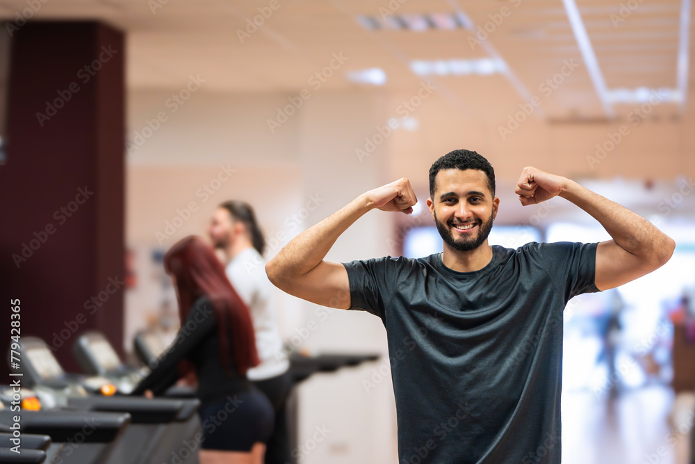 Joyful man showing off muscles with a flex at the gym, companions working out behind. Joyful man showing off muscles with a flex at the gym, companions working out behind.