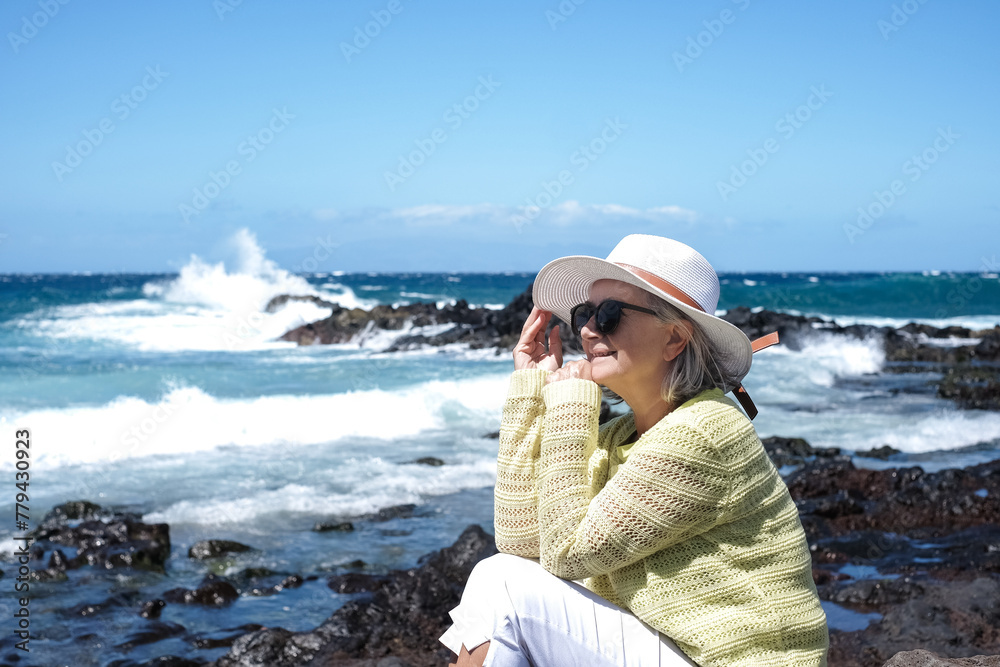 Smiling gray haired woman on a rocky beach looking at ocean waves crash on cliffs enjoying a sunny windy day, vacation or retirement lifestyle. Horizon over water, blue sky