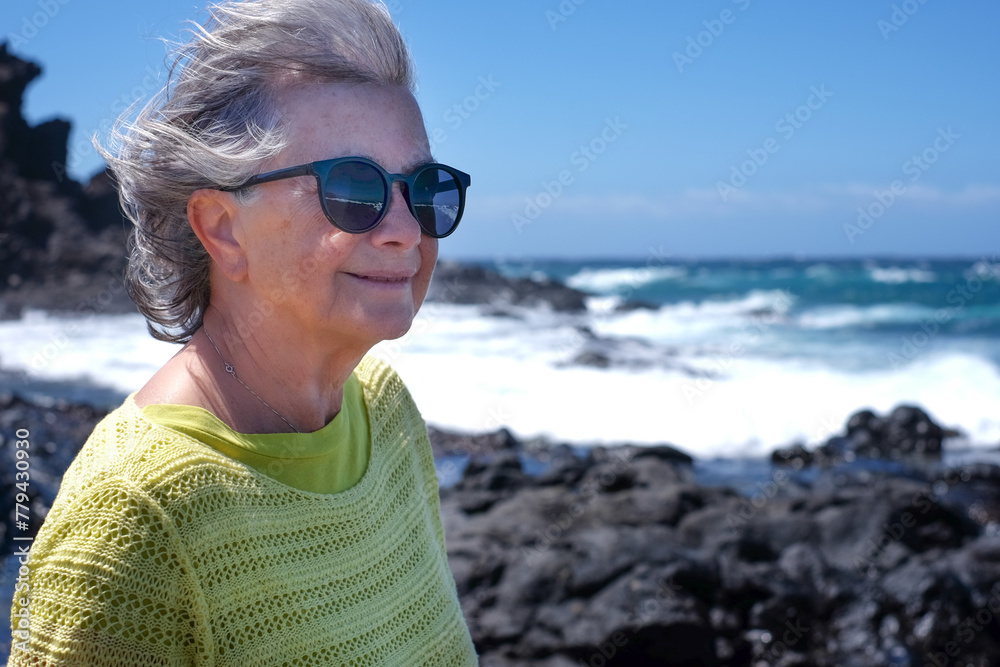 Smiling gray haired woman on a rocky beach looking at ocean waves crash on cliffs enjoying a sunny windy day, vacation or retirement lifestyle. Horizon over water, blue sky Smiling gray haired woman on a rocky beach looking at ocean waves crash on cliffs enjoying a sunny windy day, vacation or retirement lifestyle. Horizon over water, blue sky