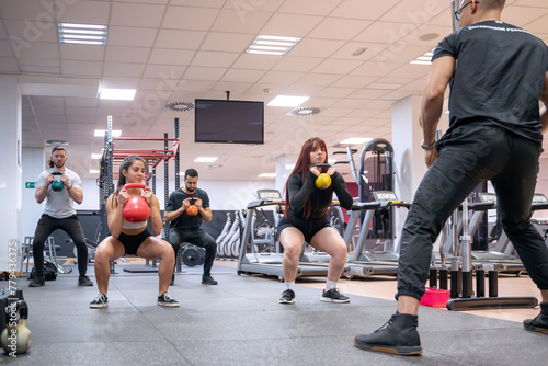 A diverse group of gym-goers engages in a kettlebell squat session with personalized coaching from a fitness instructor in a well-equipped gym.  photo