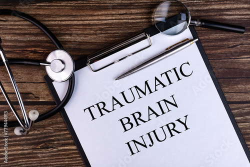 TRAUMATIC BRAIN INJURY text is written on a tablet lying on a dark table with a stethoscope and a magnifying glass. Medical concept.