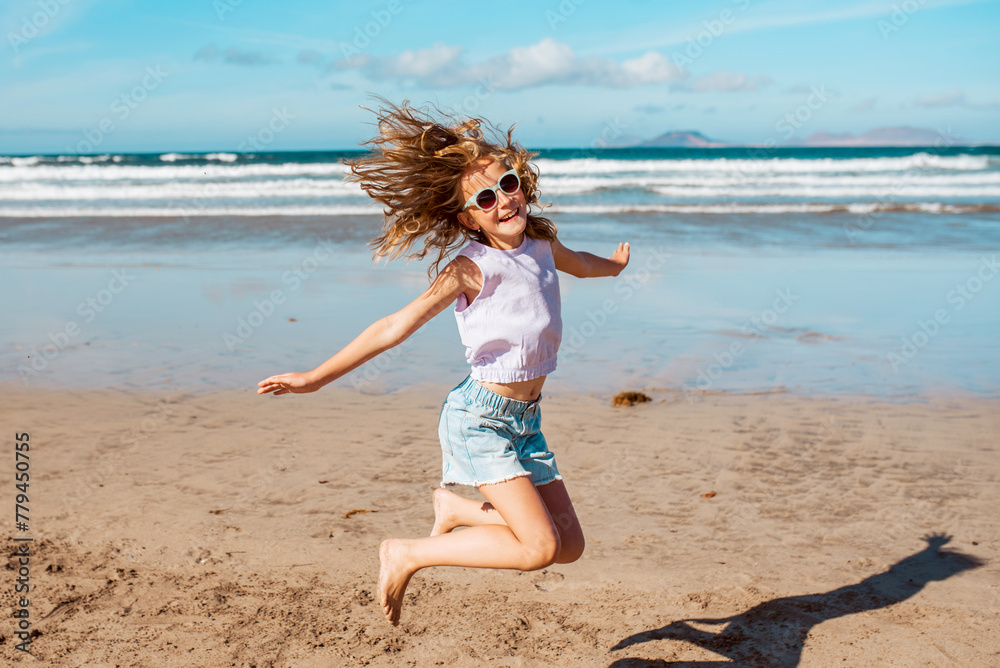 Jumping girl on beach. Smilling blonde girl enjoying sandy beach, looking at crystalline sea in Canary Islands. Concept of beach summer vacation with kids. - obrazy, fototapety, plakaty 