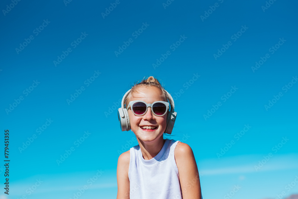 Girl with headphones on head, on beach. Smilling blonde girl enjoying sandy beach, listening music via wireless headphones. Concept of beach summer vacation with kids. Girl with headphones on head, on beach. Smilling blonde girl enjoying sandy beach, listening music via wireless headphones. Concept of beach summer vacation with kids.