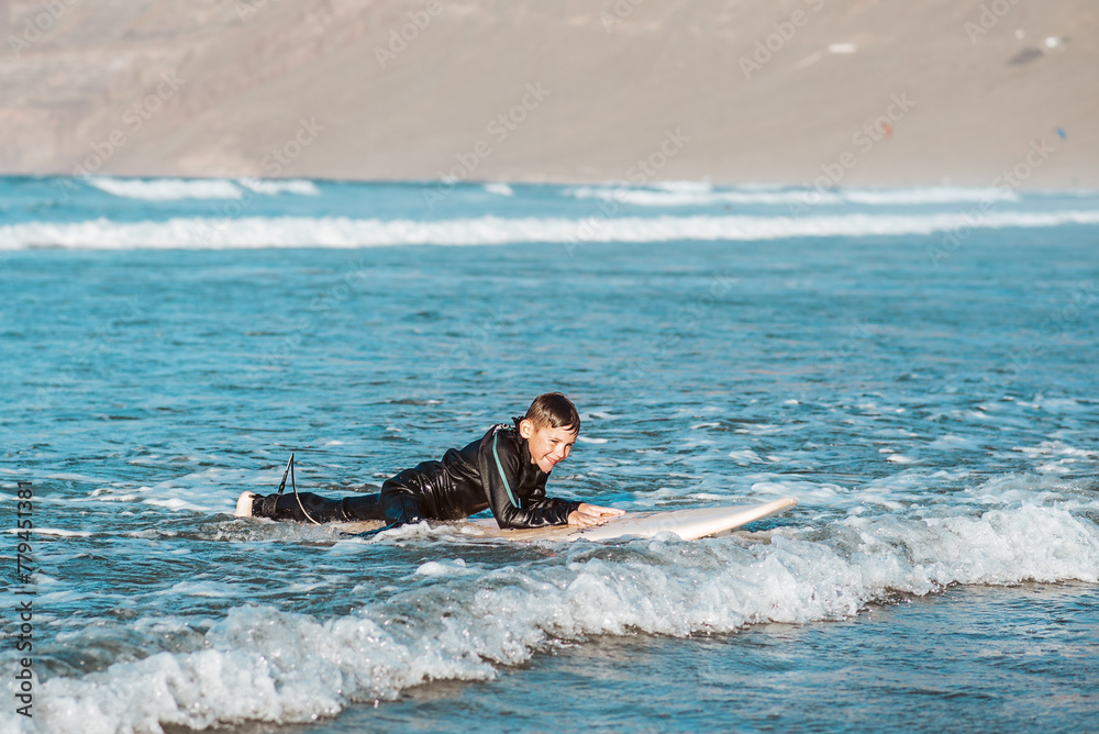 Young boy surfing in sea waves. Smilling boy in wetsuit lying on sufrboard. Concept of popular water sport for kids. Young boy surfing in sea waves. Smilling boy in wetsuit lying on sufrboard. Concept of popular water sport for kids.
