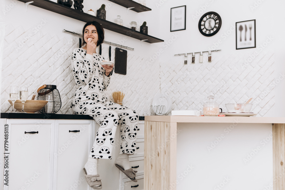 Cheerful woman standing in bright kitchen smiling eat candy, dressed in patterned pajama set. The kitchen counter arranged with ingredients, utensils
Cheerful woman standing in bright kitchen smiling eat candy, dressed in patterned pajama set. The kitchen counter arranged with ingredients, utensils
