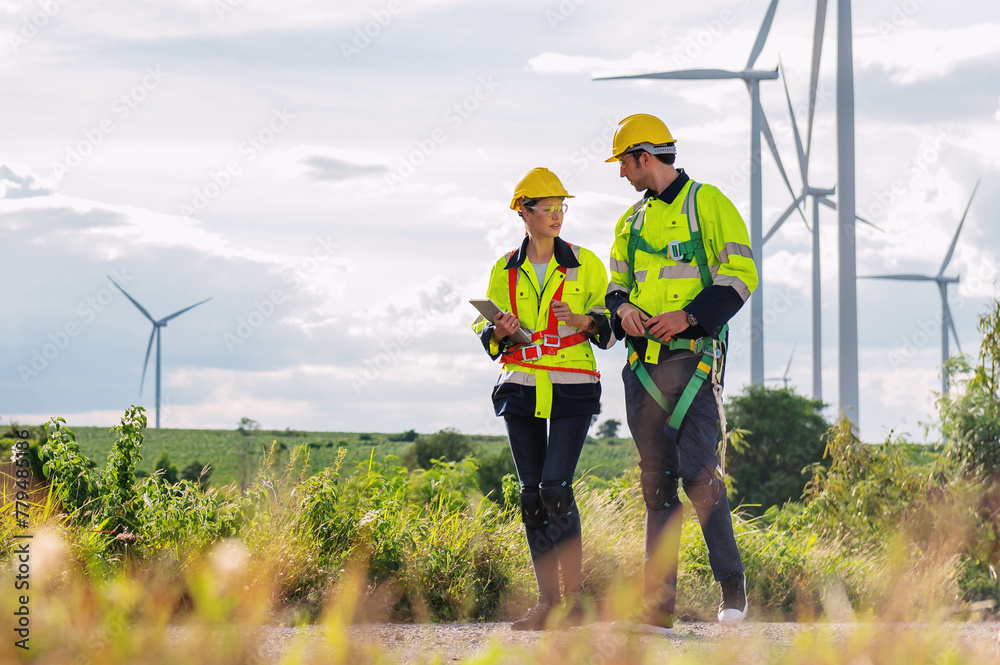 Two workers in safety gear are walking through a field of wind turbines Two workers in safety gear are walking through a field of wind turbines