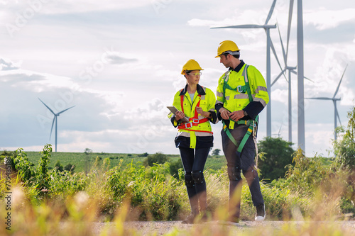Two workers in safety gear are walking through a field of wind turbines