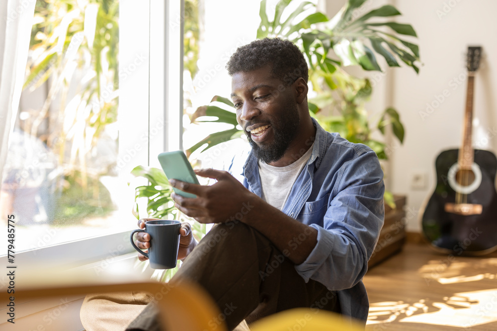 © Westend61 - Smiling non-binary person holding coffee cup and using smart phone at home
