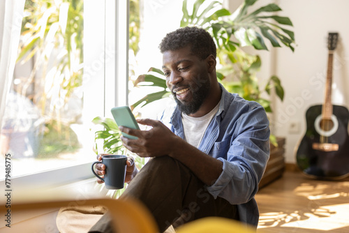 Smiling non-binary person holding coffee cup and using smart phone at home photo