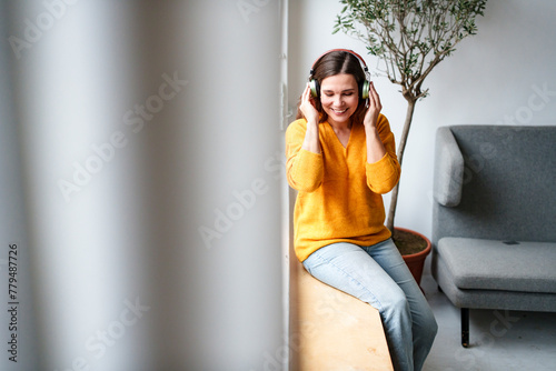 Smiling mature woman listening to music through wireless headphones sitting on window sill at home photo