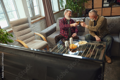 Cheers. Senior men, friends sitting on couch at home, drinking beer, eating snacks and watching online sport match translation. Concept of sport, championship, leisure and entertainment