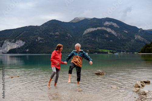 Active elderly couple hiking together in spring mountains, walking in cold water in mountain lake. Senior tourists enjoying nature.