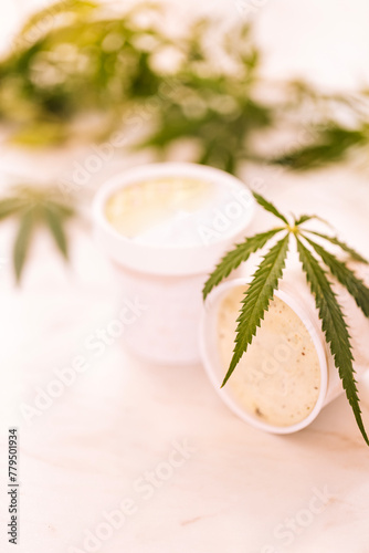 A hemp leaf is lying on a mock-up jars of ice cream on a white marble table. Green cannabis leaves on the background.