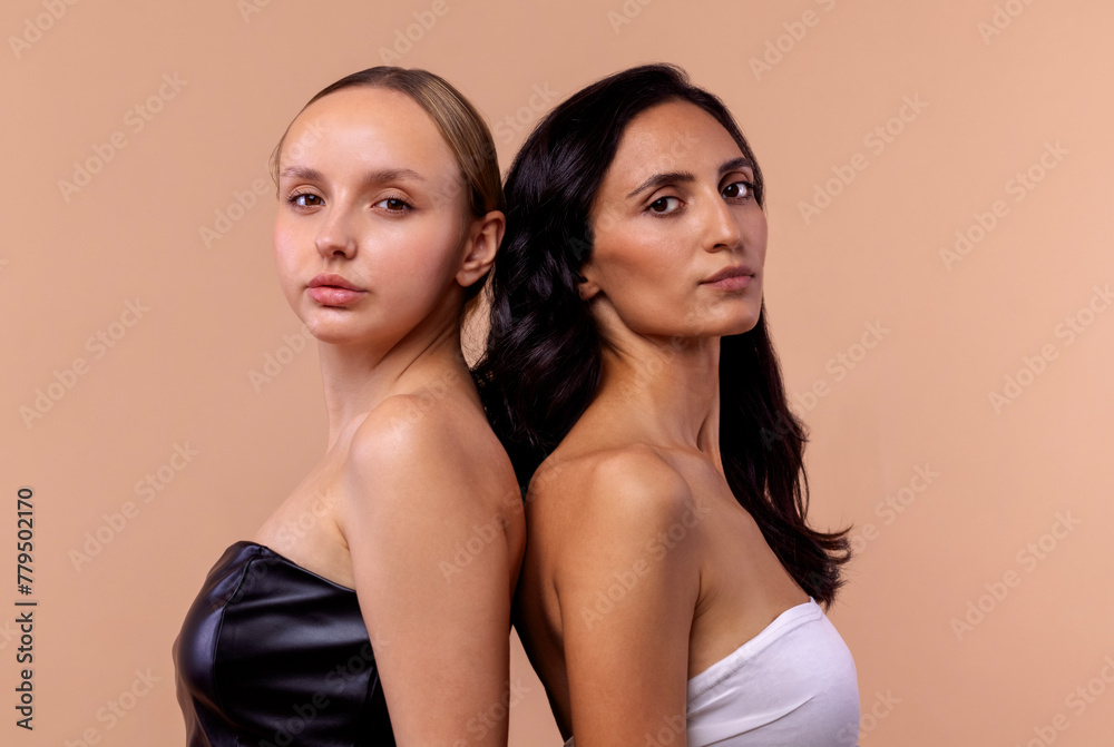 Two young diverse women wearing tops looking confident at camera while posing together on beige background. Charming models with natural makeup and perfect skin. Two young diverse women wearing tops looking confident at camera while posing together on beige background. Charming models with natural makeup and perfect skin.