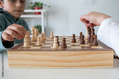 Two children playing a game of chess. One of the boys is wearing a blue hoodie, the boy is about to make a move. The game is played on a wooden board with black and white squares. 
