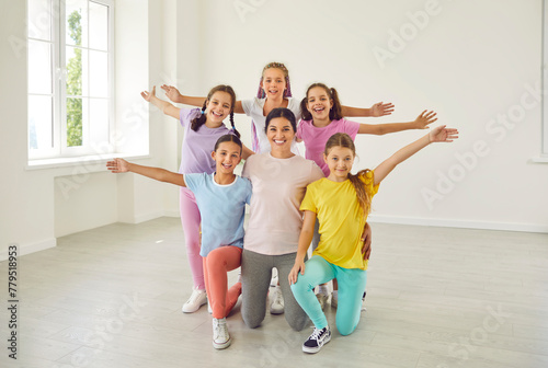 Portrait of cheerful happy active little girls sitting on the floor with their smiling friendly choreographer woman in choreography class, posing and looking at camera after dance workout.