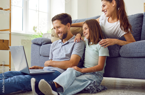 Happy young family of three with child girl sitting on the floor at home using laptop together having video call talking online. Smiling parents with daughter watching funny cartoon or movie.