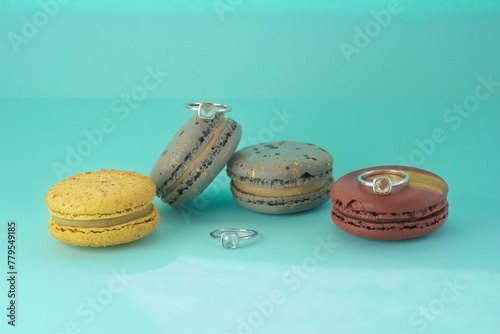 close-up of set of three elegant engagement silver rings with a gemstones between multicolor macaronies cakes on an azure background