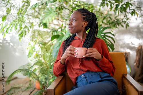 Relaxed dreaming african american woman holding cup of coffee sitting on chair in urban jungle interior lookin aside window. Pleased thoughtful black female resting on armchair surround houseplants. photo