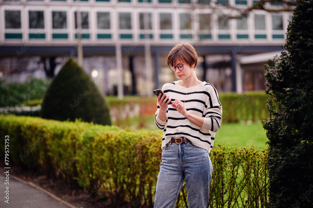 A young woman in casual attire is engaged with her smartphone in an urban garden, showcasing a moment of modern connectivity in a natural setting.