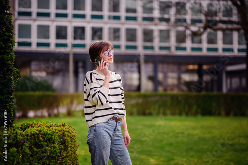 A lively chat unfolds as a stylish woman in a striped sweater engages in a phone conversation, her animated expression adding life to the serene urban park setting.