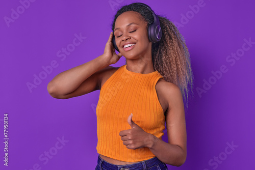 Young cheerful emotional African American woman in wireless headphones shows thumbs up and closes eyes with pleasure hearing music track of favorite singer stands in purple studio. Podcast, audio book