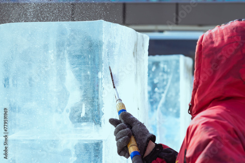A sculptor processes a large ice cube with a sharp hand tool. Ice dust and ice fragments in the air. Ice sculpture festival in the city.