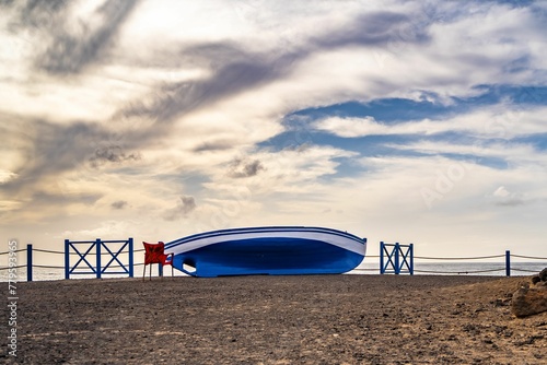 Beautiful shot of a blue boat laid ashore near a red chair on Fuerteventura island in Spain © Wirestock