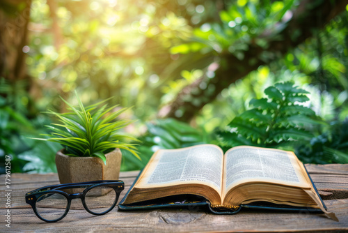 An open book with glasses next to it on a wooden table with a green vase and green vegetation in the background, outdoor reading theme
 photo