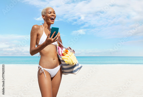 Happy woman at the beach side wearing bikini holding a beach bag and using mobile phone in a sunny day with blue sky. Concept of summer beach holiday, shopping online, booking travel and resort