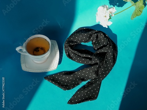 A composition of a cup of herbal tea with a saucer and a silk dark blue polka dot neckerchief and a white flower on a bright blue background with rays of sunlight and shadows     