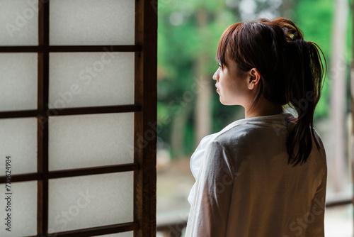 A contemplative Japanese woman stands at the threshold of a traditional wooden veranda, gazing out at the serene green landscape.