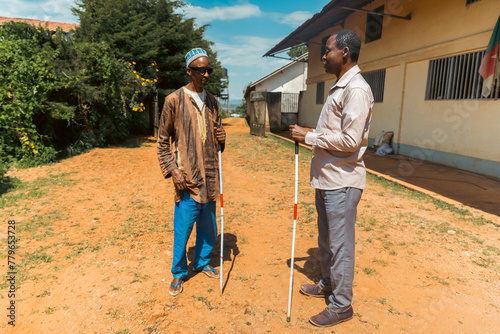 Two men are standing in a dirt field, one of them holding a cane