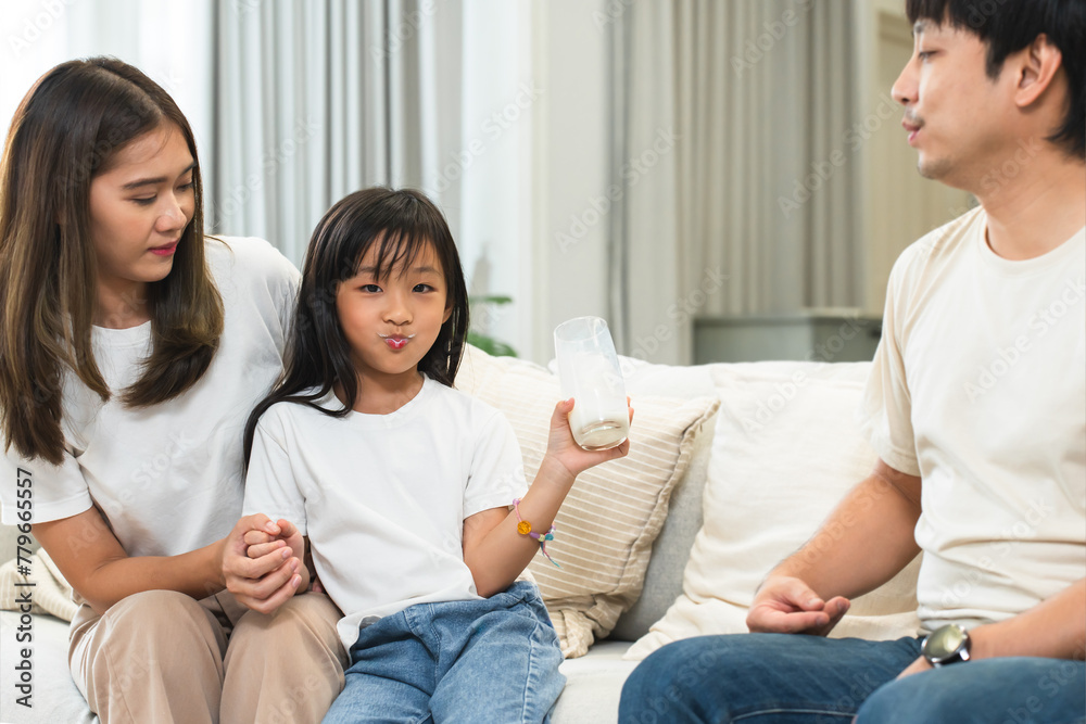 Happy Asian family, father and mother looking at little 6 year old daughter drinking, holding glass of milk with milk stained mouth, looking at camera, sitting on sofa at home. Selective focus on kid