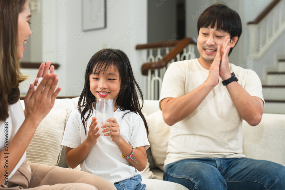 Happy Asian family, mother and father clapping hands cheering their little 6 year old daughter drinking milk from glass with smiling face in morning, sitting on sofa at home. Selective focus on child Happy Asian family, mother and father clapping hands cheering their little 6 year old daughter drinking milk from glass with smiling face in morning, sitting on sofa at home. Selective focus on child