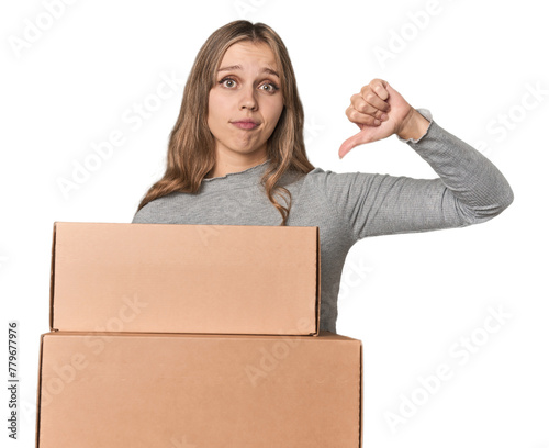 Blonde woman with cardboard boxes moving on studio backdrop showing a dislike gesture, thumbs down. Disagreement concept.