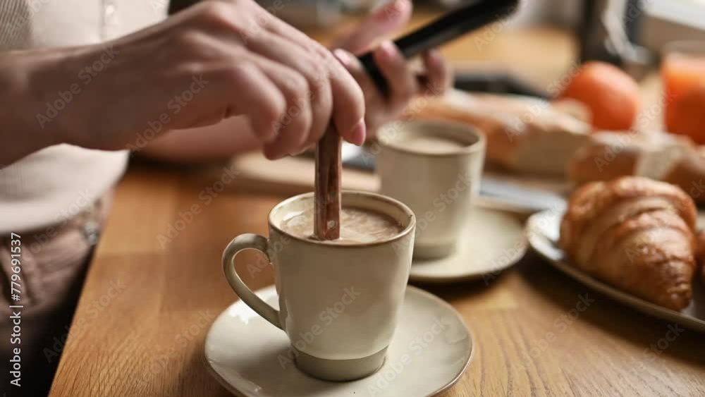 Girl hand stirs coffee cappuccino in cup with spoon and looking smartphone screen. Woman with creamy espresso and mobile phone at kitchen Girl hand stirs coffee cappuccino in cup with spoon and looking smartphone screen. Woman with creamy espresso and mobile phone at kitchen