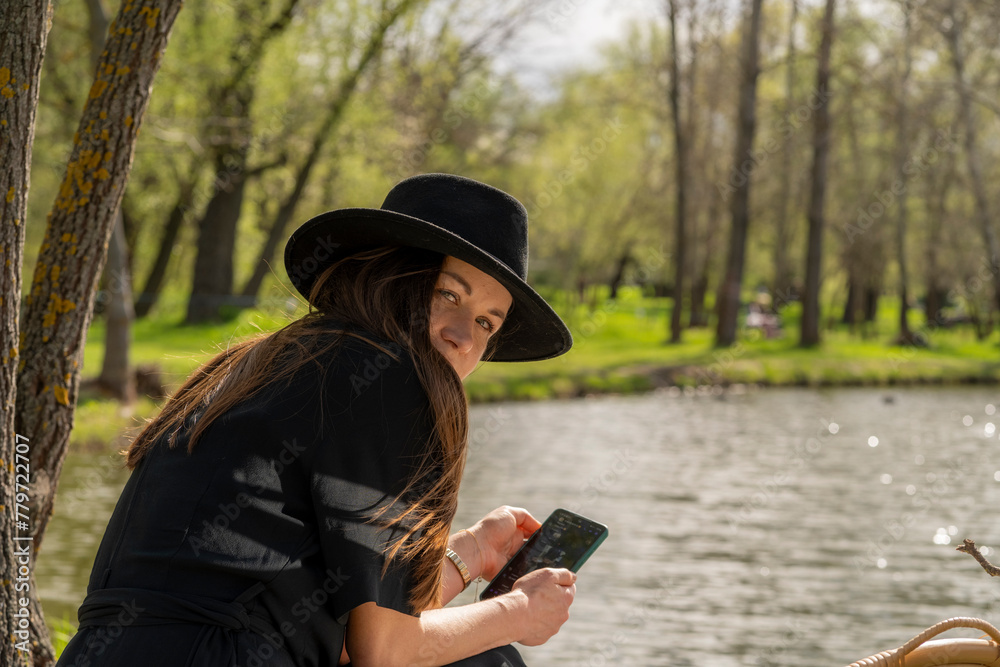 Girl in a black hat flirtatiously looks at the camera holding a phone in her hands on the lake shore